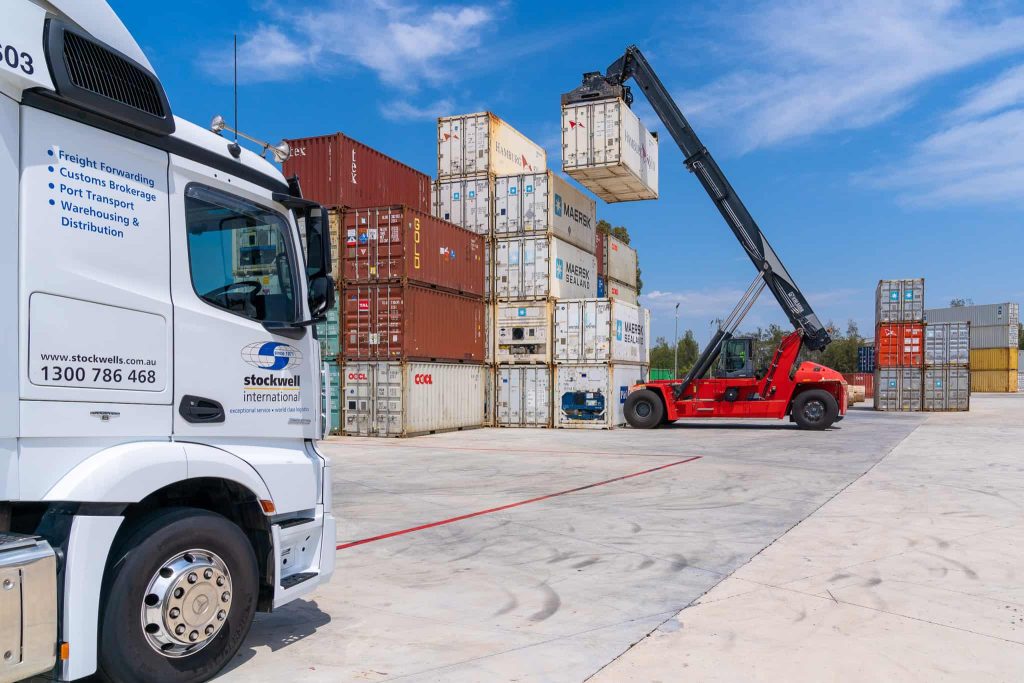 port transport - crane moving shipping container with freight truck waiting in the foreground